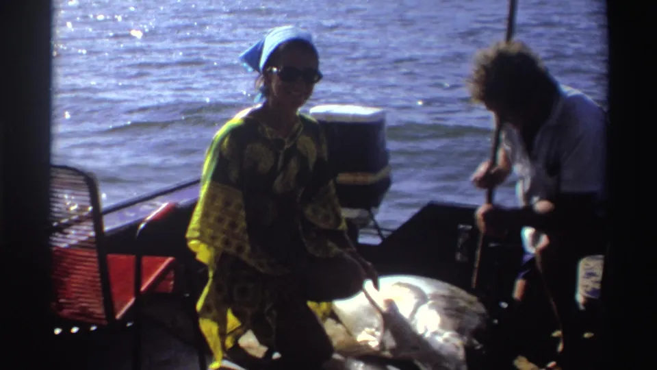 1969 Nigeria home-movie frame with a person in a blue headscarf near the fish and another person seated by the outboard motor.