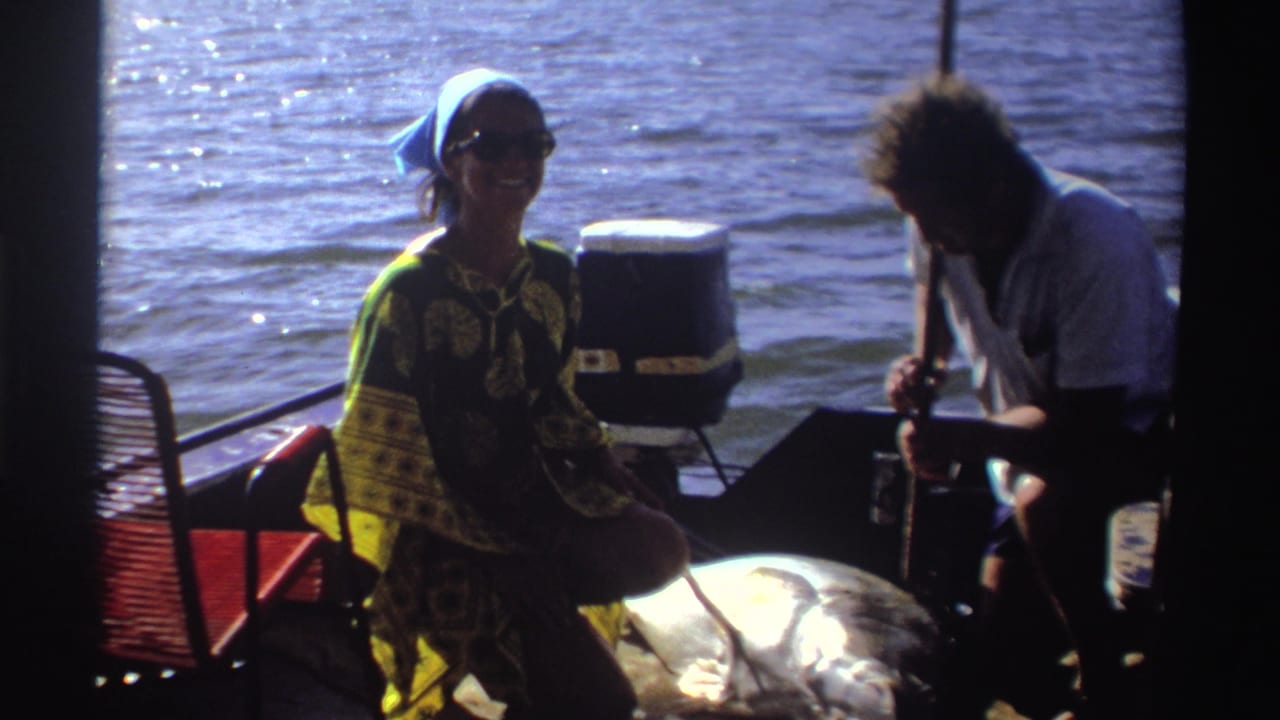 Woman in patterned dress smiles on a small boat as a man tends to a large fish beside her.