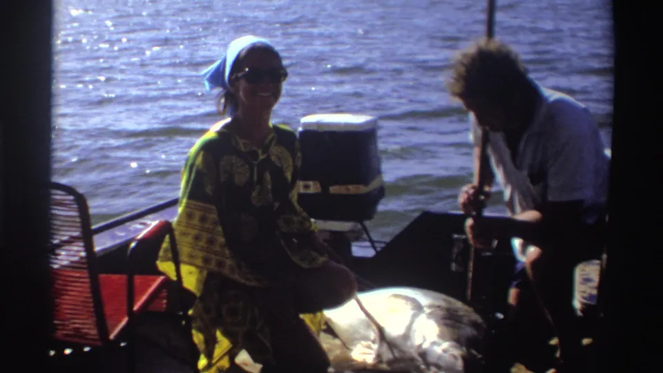 In 1969 Nigeria, a woman in a blue headscarf smiles toward the camera while a man leans over a large fish on a small boat.