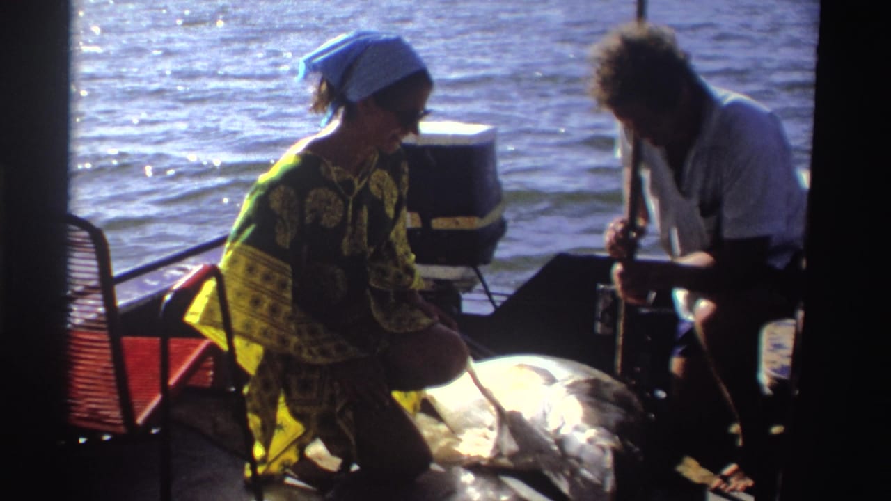 Woman turns toward a man seated near a large fish on a sunlit boat with water behind them.