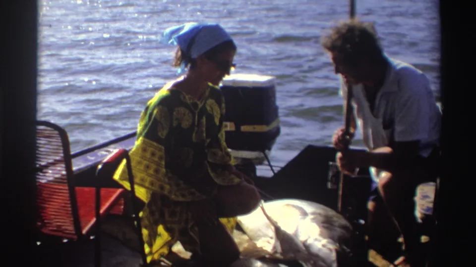 The 1969 Nigeria boating scene shows the woman looking down while the man works near the large fish on the deck.