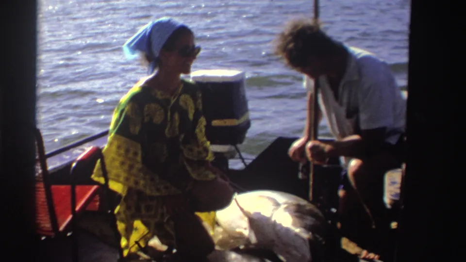 In this Nigeria 1969 8mm frame, the woman sits in profile as the man leans over near the large fish on the boat.