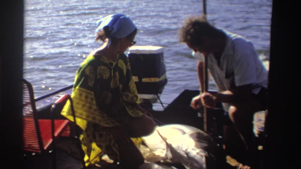 1969 Nigeria 8mm frame of a woman in a green-yellow kaftan on a small boat as a man leans over near a large fish.
