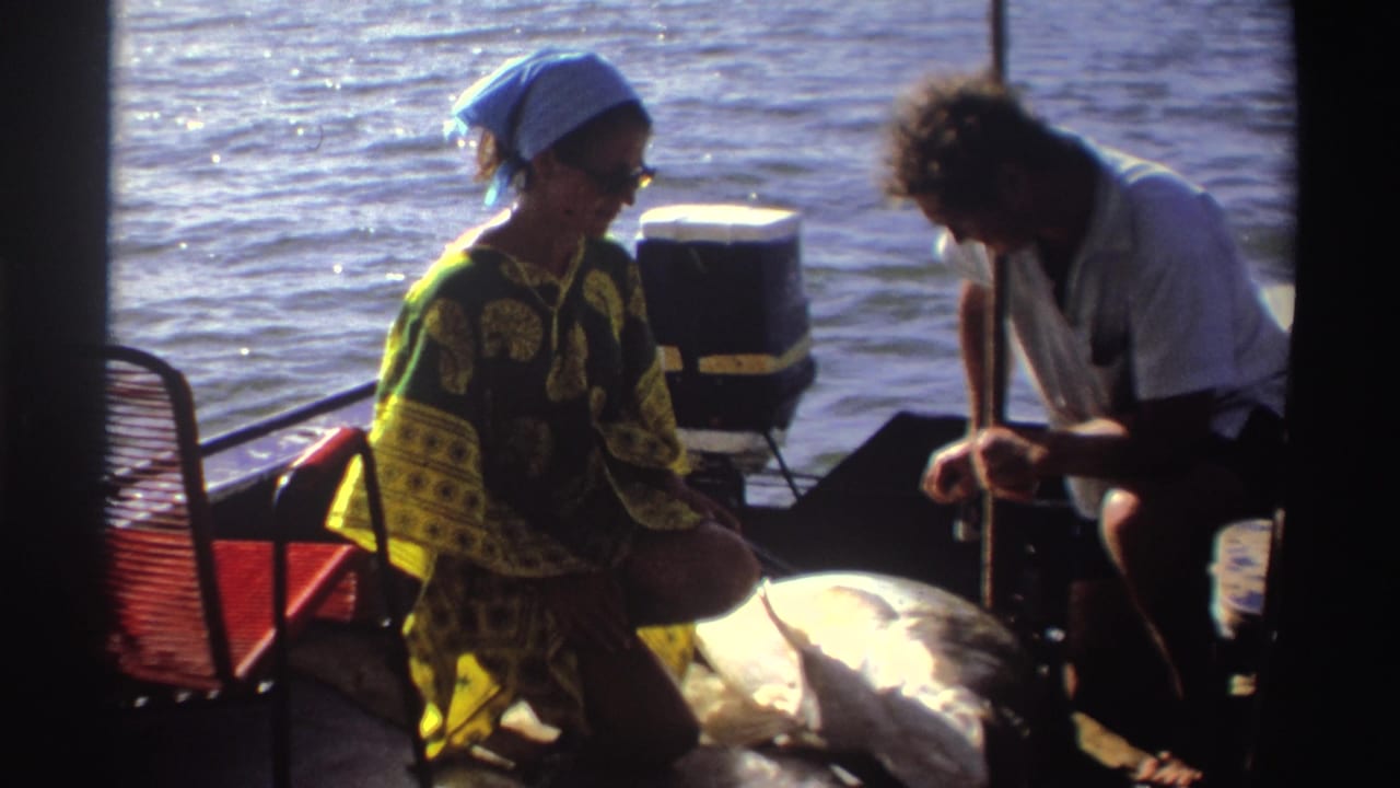 Woman seated on a boat faces a man leaning over fishing gear near a large fish.