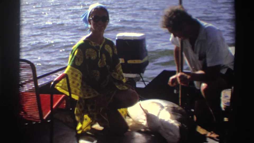 1969 Nigeria 8mm frame of a woman in sunglasses and a blue headscarf smiling from a small fishing boat beside a large fish.