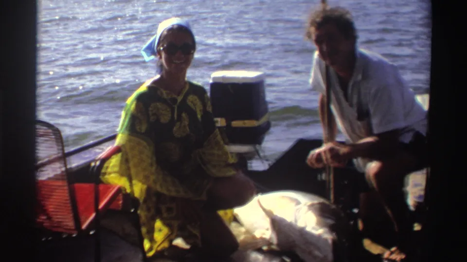 1969 Nigeria home-movie frame with two adults on a small fishing boat, the woman smiling and a large fish visible between them.