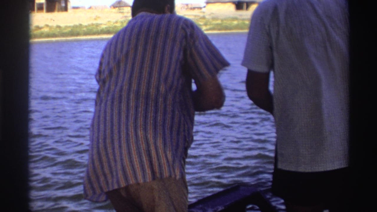1969 Nigeria home-movie frame showing two adults from behind on a river boat, with rippling water and a shoreline in the background.