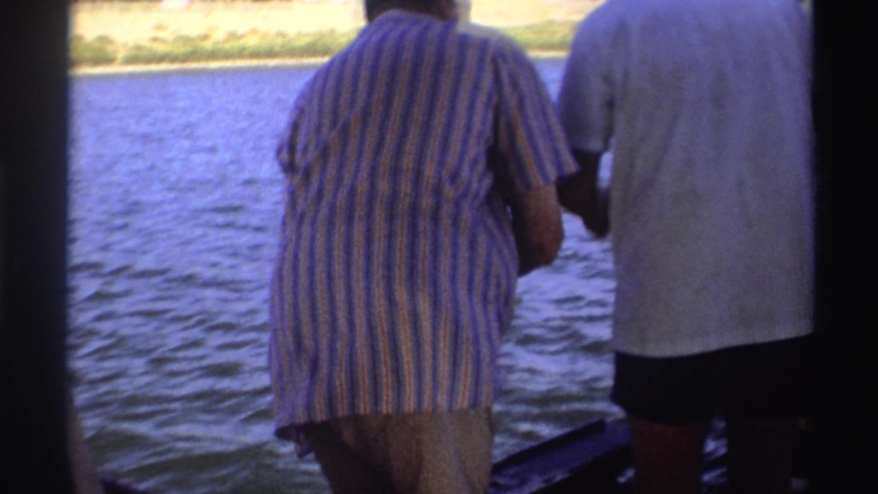 1969 Nigeria frame showing two adults standing at the side of a small boat with river water and distant shoreline houses visible.