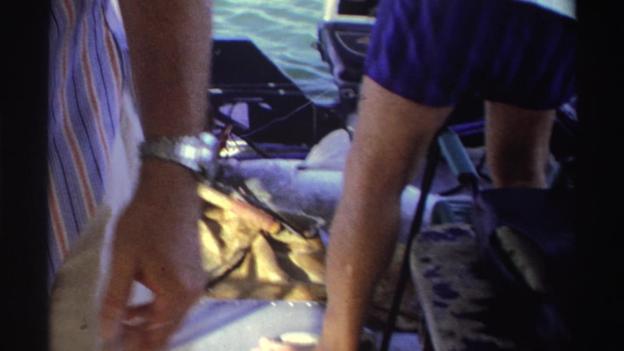 1969 Nigeria home-movie frame showing adults on a boat with a large fish on deck and water beyond the rail.