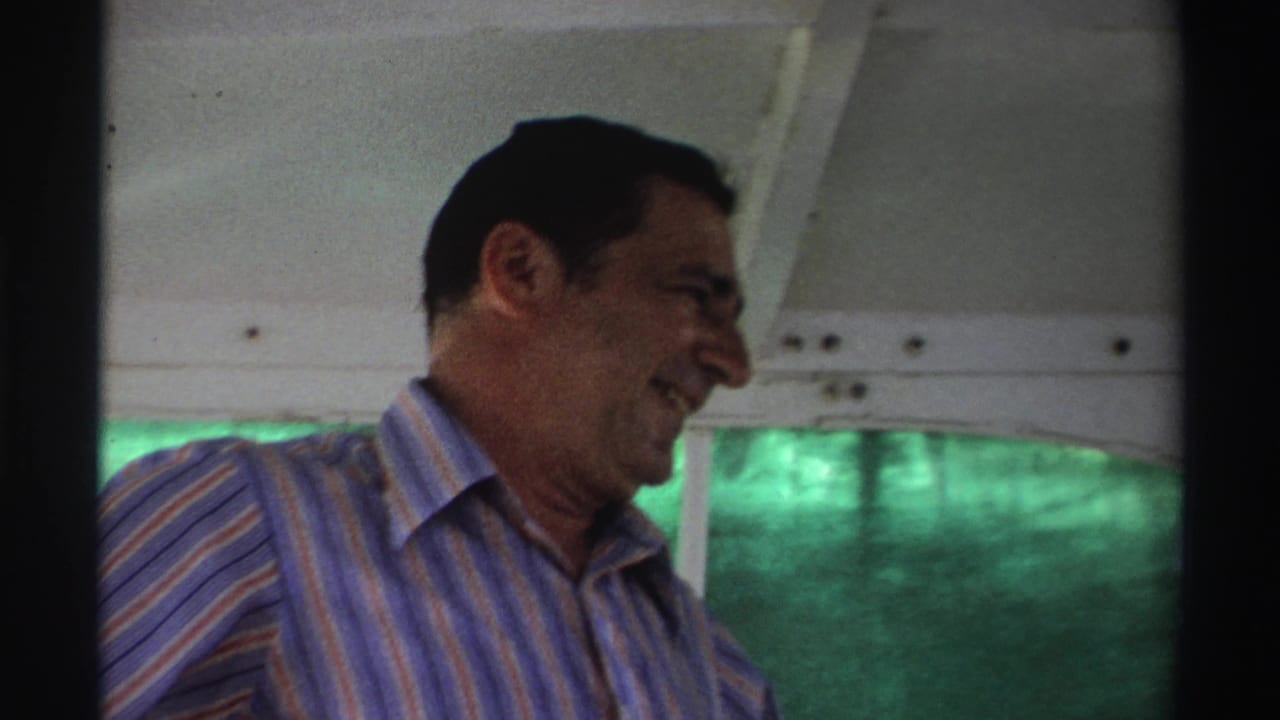 In Nigeria, 1969, a middle-aged man in a blue and yellow striped shirt smiles while seated or leaning inside a boat.