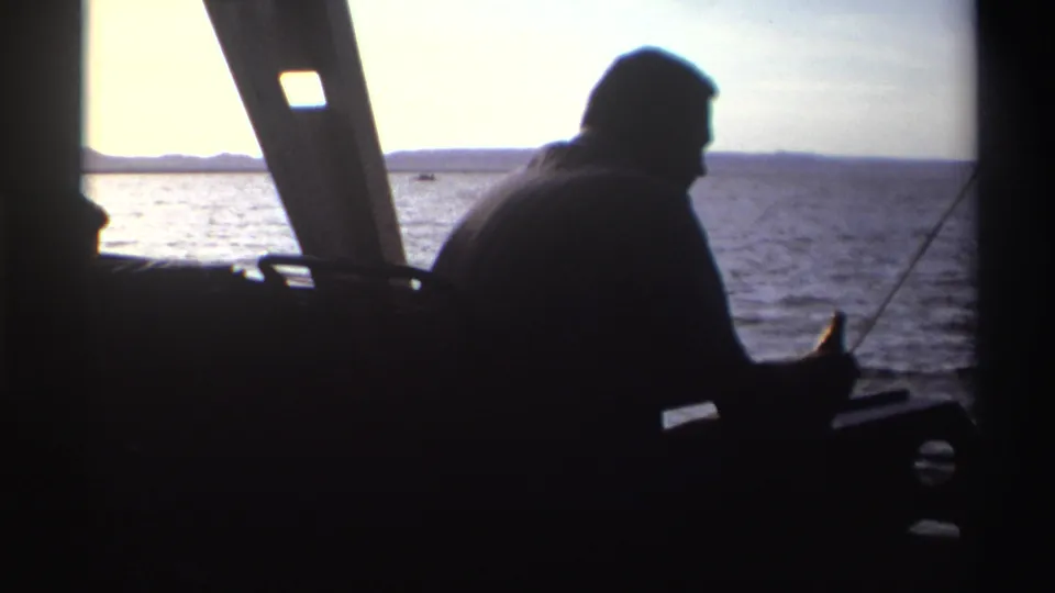 A silhouetted adult boater is seen from inside a small vessel, with bright water and distant land beyond the cabin frame.