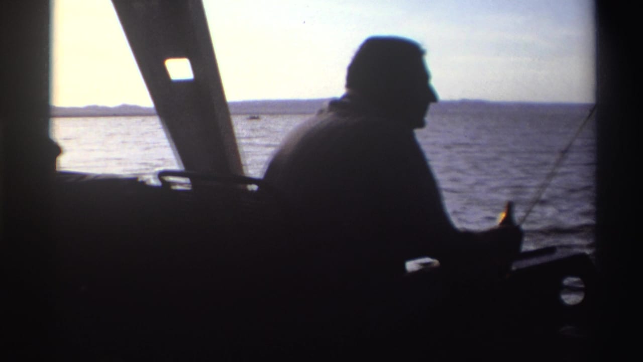 The boater remains in dark profile inside the cabin, framed against calm water and a pale sky in the 1969 Nigeria footage.