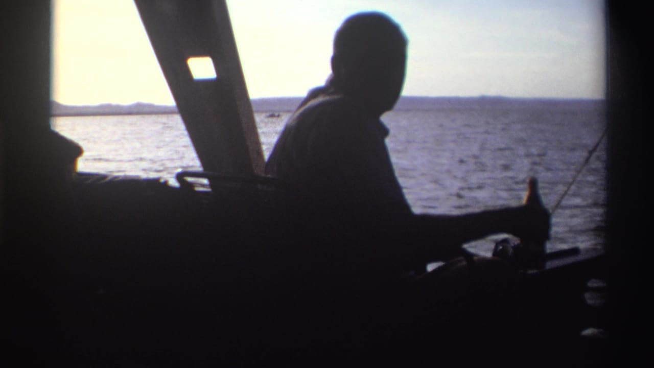 Silhouetted man sits inside a boat holding a bottle beside open water.