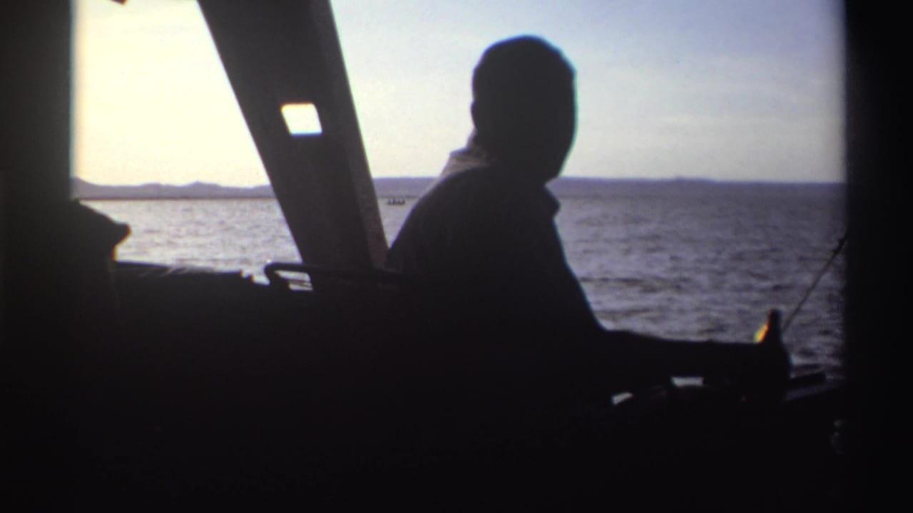Silhouetted adult passenger in a boat in Nigeria, 1969, with a bottle near his hand and distant land across the water.