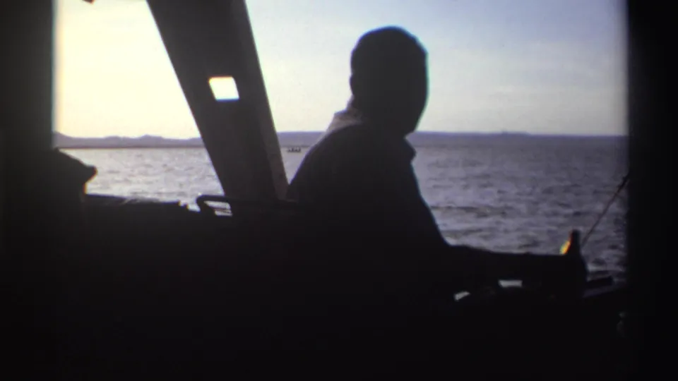 Silhouetted adult passenger in a boat in Nigeria, 1969, with a bottle near his hand and distant land across the water.