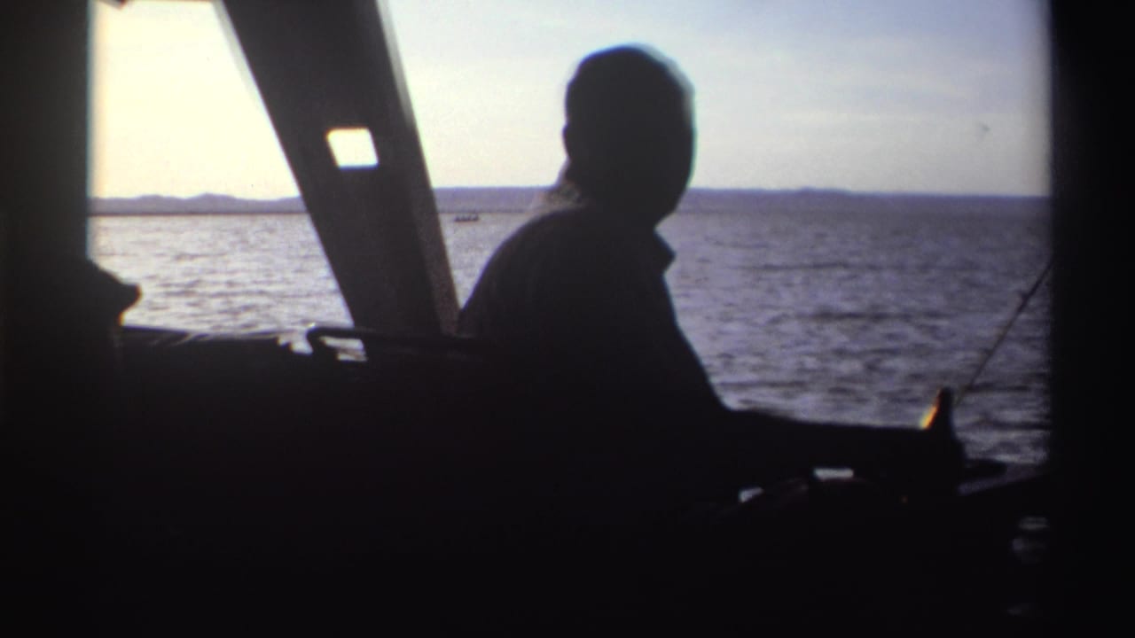 Home-movie frame from 1969 Nigeria showing a backlit seated man in a boat, facing water and a low distant shoreline.