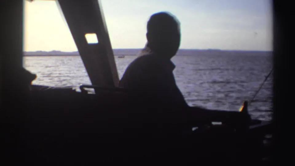 Home-movie frame from 1969 Nigeria showing a backlit seated man in a boat, facing water and a low distant shoreline.