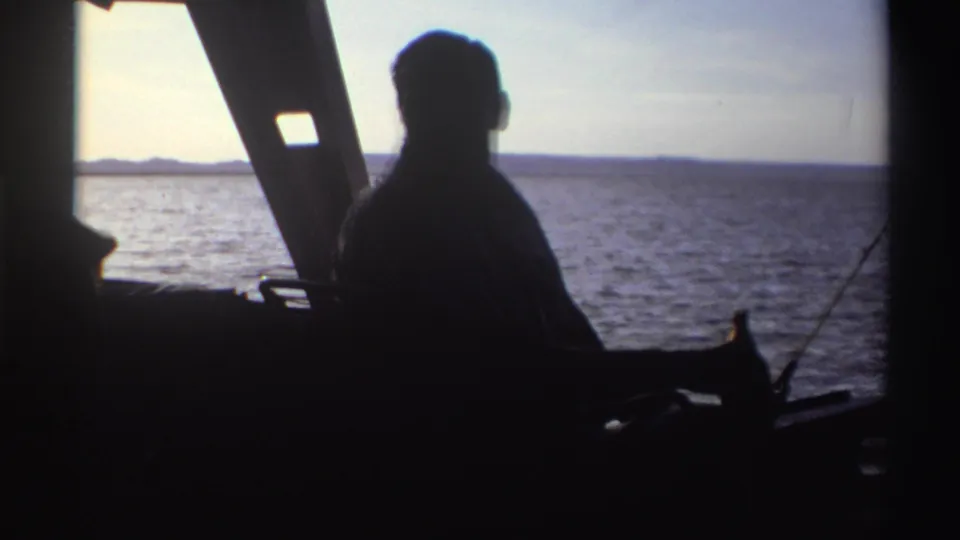 1969 Nigeria 8mm still of a man seen in silhouette from inside a boat, looking toward open water and distant land.