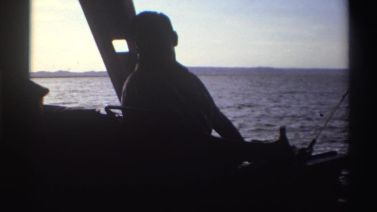Silhouetted man seated inside a boat with open water and distant shoreline behind him.