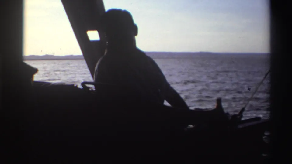 In 1969 Nigeria home-movie footage, a silhouetted man sits on a boat against bright water and a low horizon.