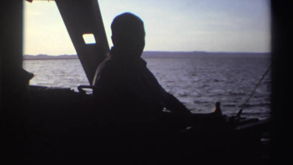 The silhouetted man remains seated on the boat, partly turned toward the camera with water and horizon behind him.