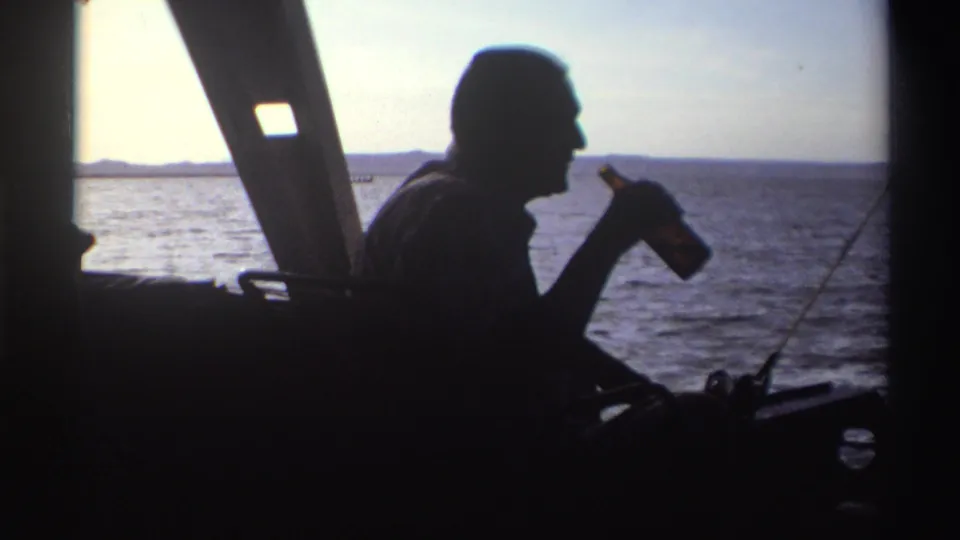 The man raises a bottle near his mouth while seated in silhouette on the boat, with Nigerian waters visible beyond.