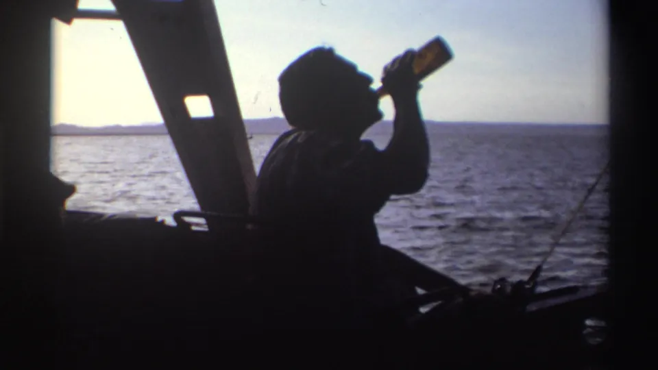 A silhouetted man drinks from a bottle aboard the boat, framed by bright water, sky, and a dark structural support.
