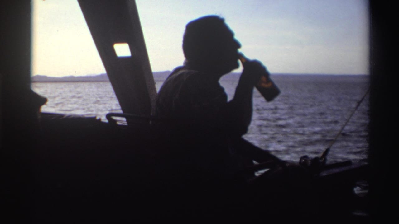 Silhouetted man on a boat drinks from a brown bottle beside the ocean.