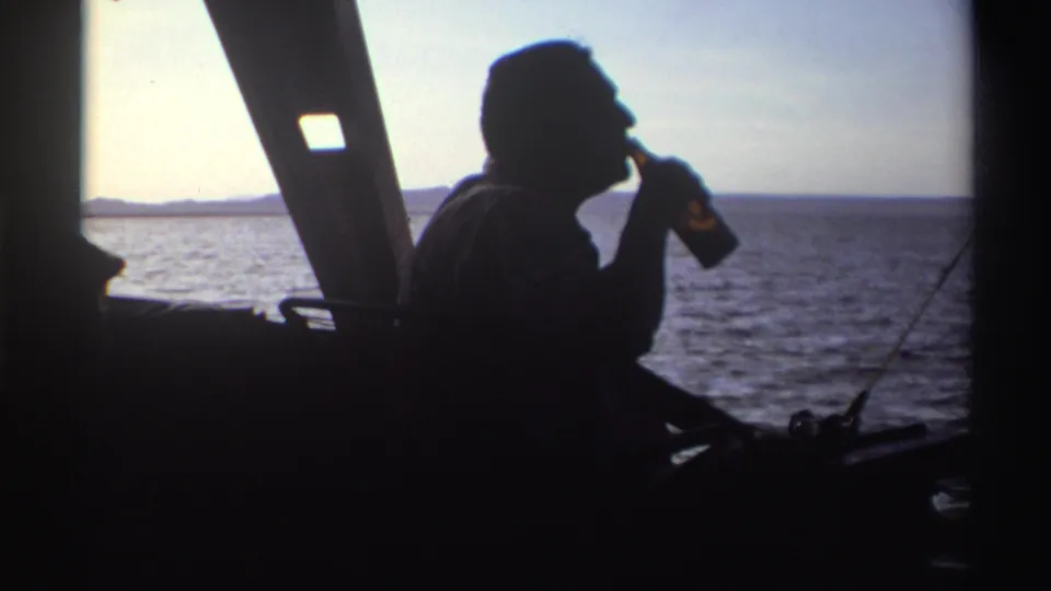In Nigeria 1969 home-movie footage, a silhouetted man drinks from a bottle on a boat with open water behind him.