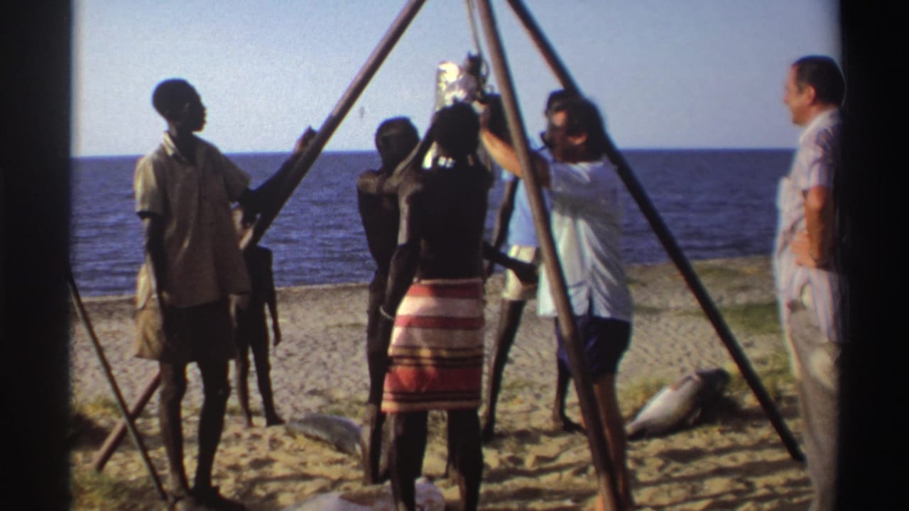 Nigeria, 1969: adults on a sandy shoreline cluster around a hoisted fish, with tripod poles framing the scene.
