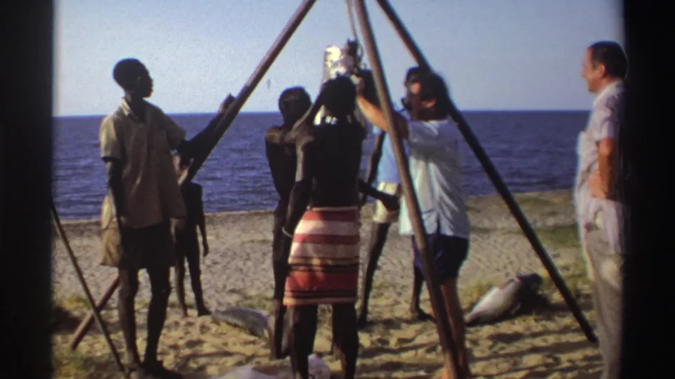 Nigeria, 1969: adults on a sandy shoreline cluster around a hoisted fish, with tripod poles framing the scene.