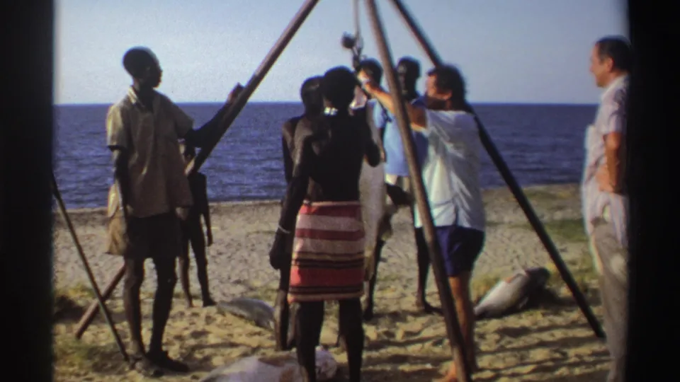 Nigeria, 1969: people on the beach inspect or adjust a large fish hanging from a wooden hoist near the water.