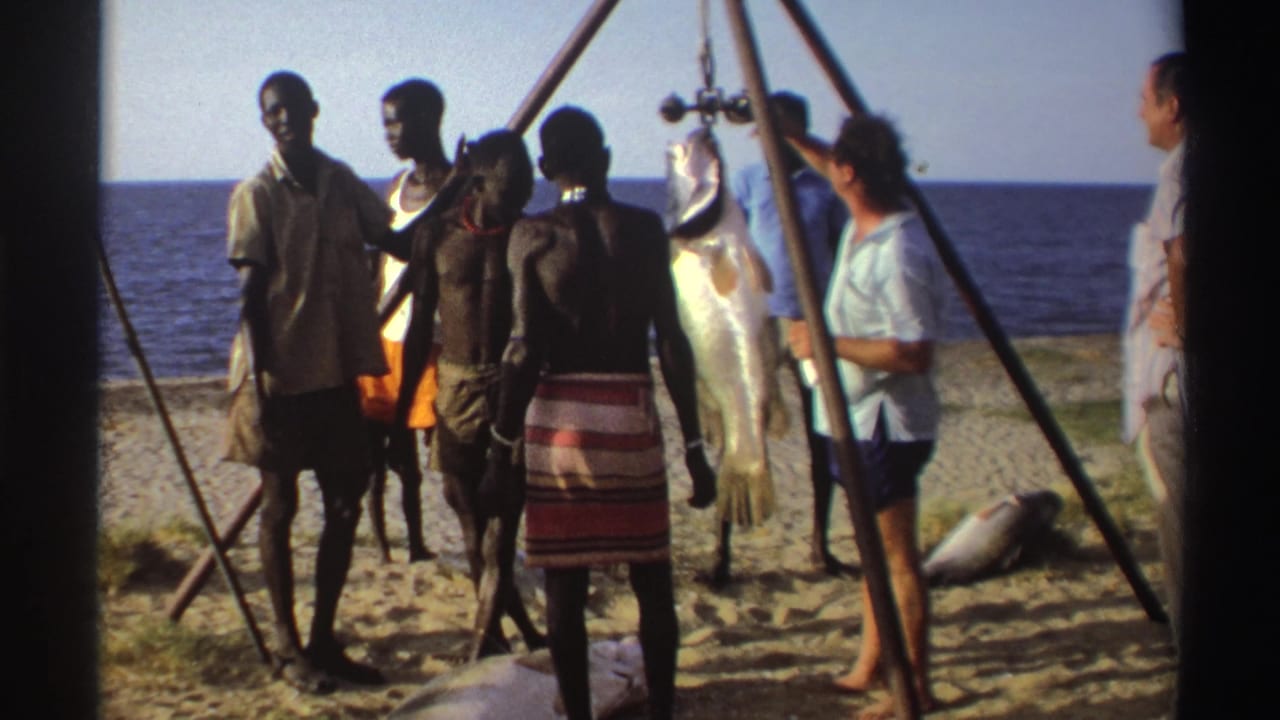 Nigeria, 1969: men in mixed local and casual clothing stand around a large fish hanging from a beachside tripod scale.