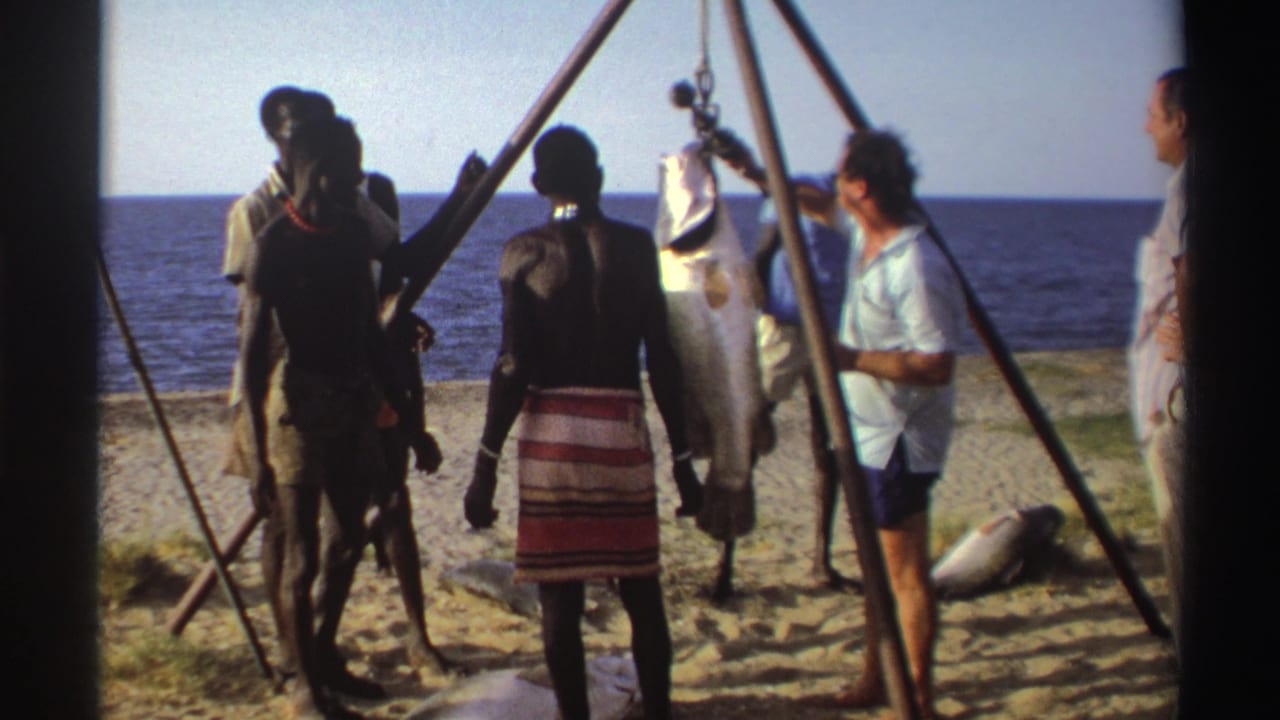 Nigeria, 1969: men move around a suspended fish under a pole tripod, filmed from a home-movie viewpoint on the beach.