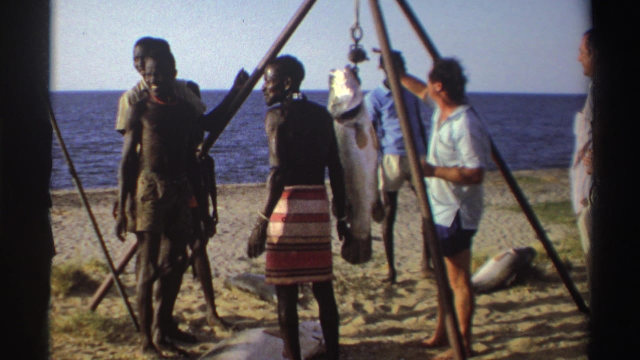 Nigeria, 1969: a man in a red and white striped wrap stands near the hanging fish as other men observe beside the ocean.