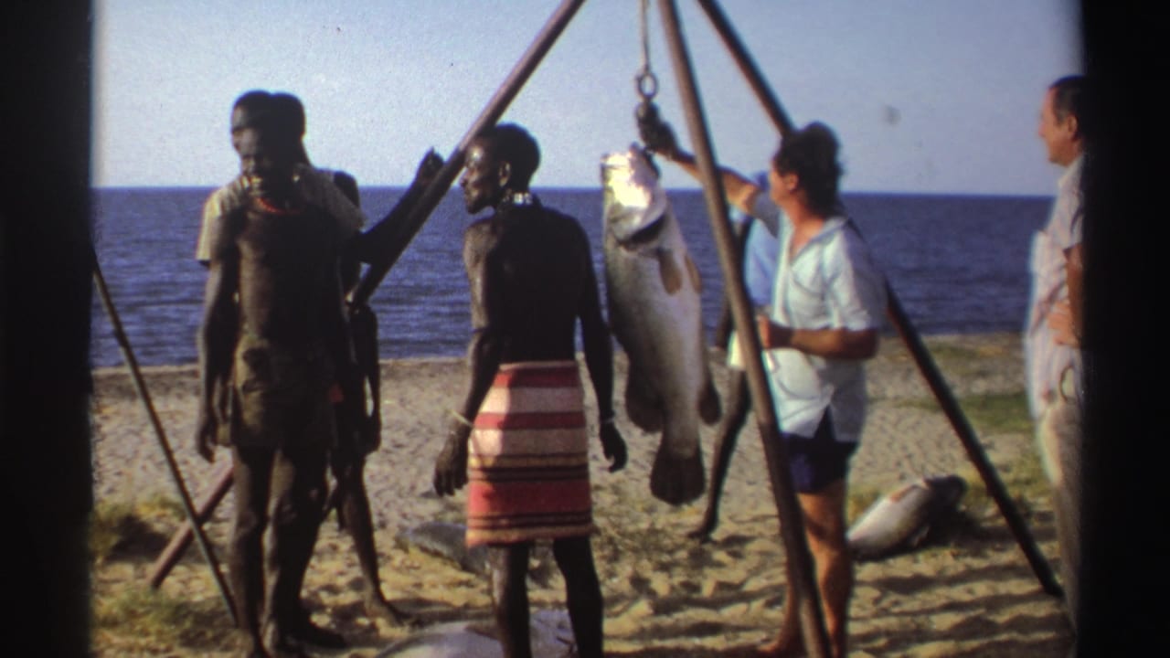 Nigeria 1969 8mm frame showing adults on a beach near the ocean gathered around a large fish suspended from a wooden tripod.