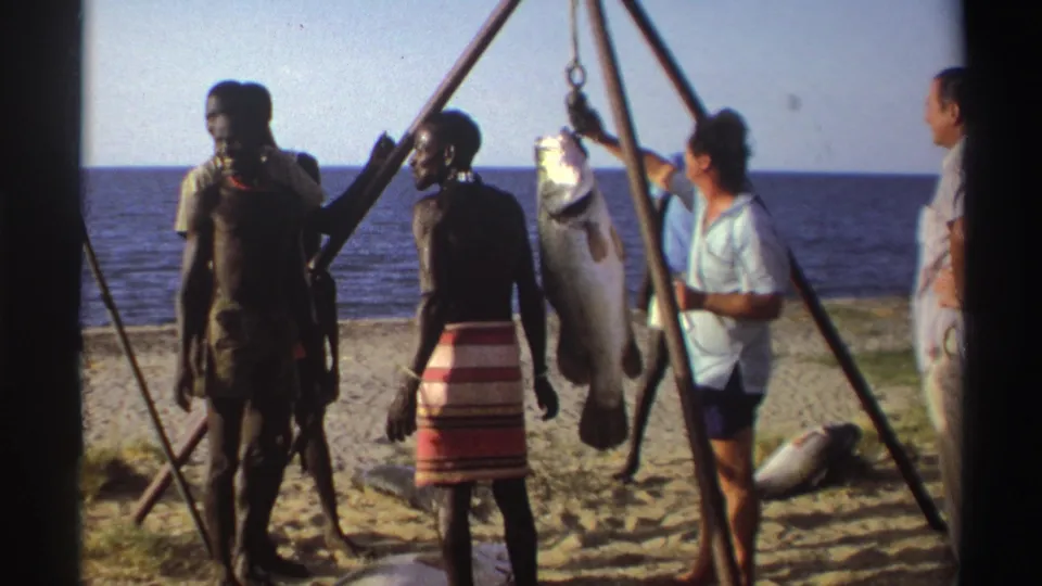 Nigeria 1969 8mm frame showing adults on a beach near the ocean gathered around a large fish suspended from a wooden tripod.