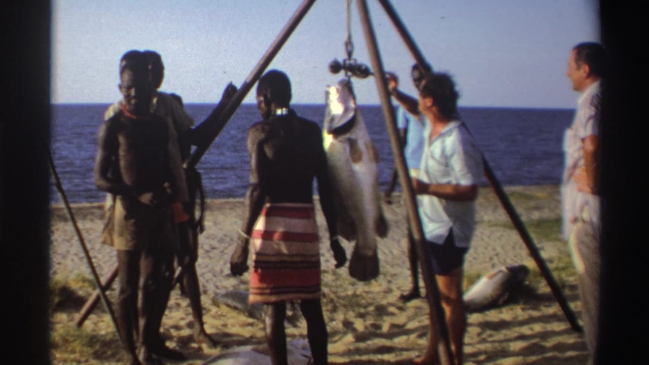Nigeria 1969 8mm frame with several adults beside a beach tripod holding a large suspended fish, ocean visible behind them.