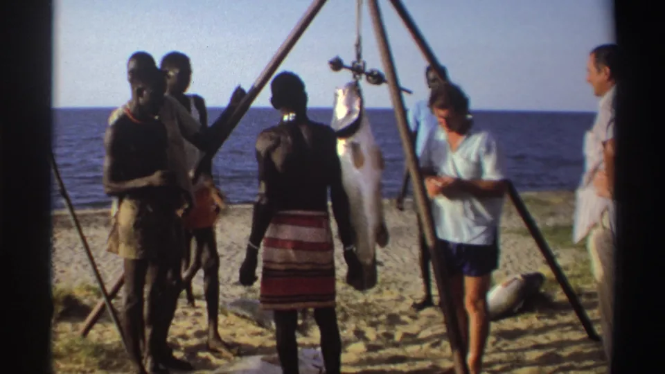 Nigeria 1969 8mm frame showing adults near the shoreline, including a large fish hanging from a wooden tripod on the sand.