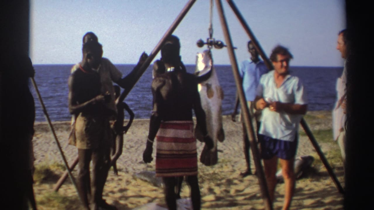 Nigeria 1969 8mm frame of adults on a sandy shore around a large hanging fish, with one person in a light shirt moving toward the camera.