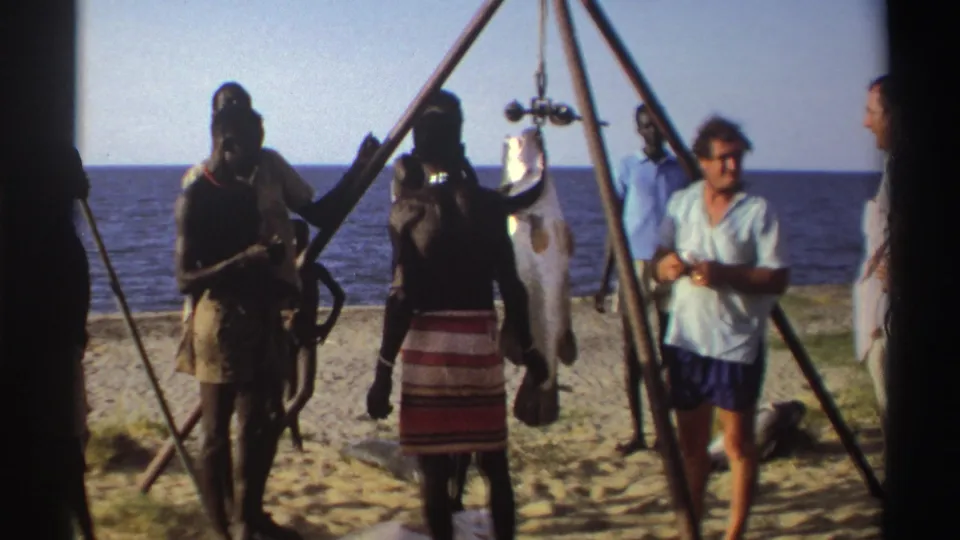 Nigeria 1969 8mm frame of adults on a sandy shore around a large hanging fish, with one person in a light shirt moving toward the camera.