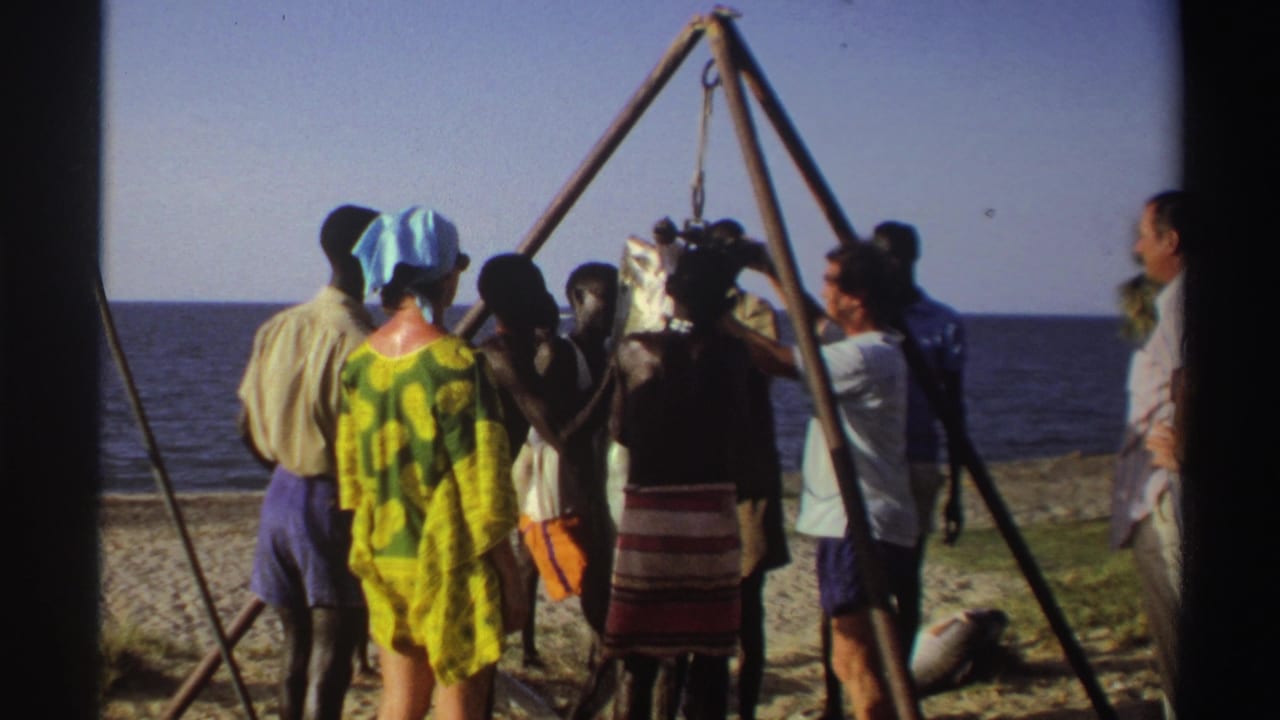 Nigeria 1969 8mm frame showing adults on a sandy beach watching a large fish hoisted from a wooden tripod near the ocean.