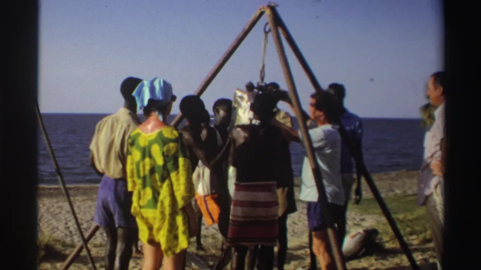 Nigeria 1969 8mm frame showing adults on a sandy beach watching a large fish hoisted from a wooden tripod near the ocean.