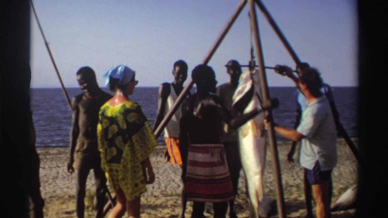 Adults in a 1969 Nigeria beach scene stand near a large catch suspended from a tripod, with the sea in the background.