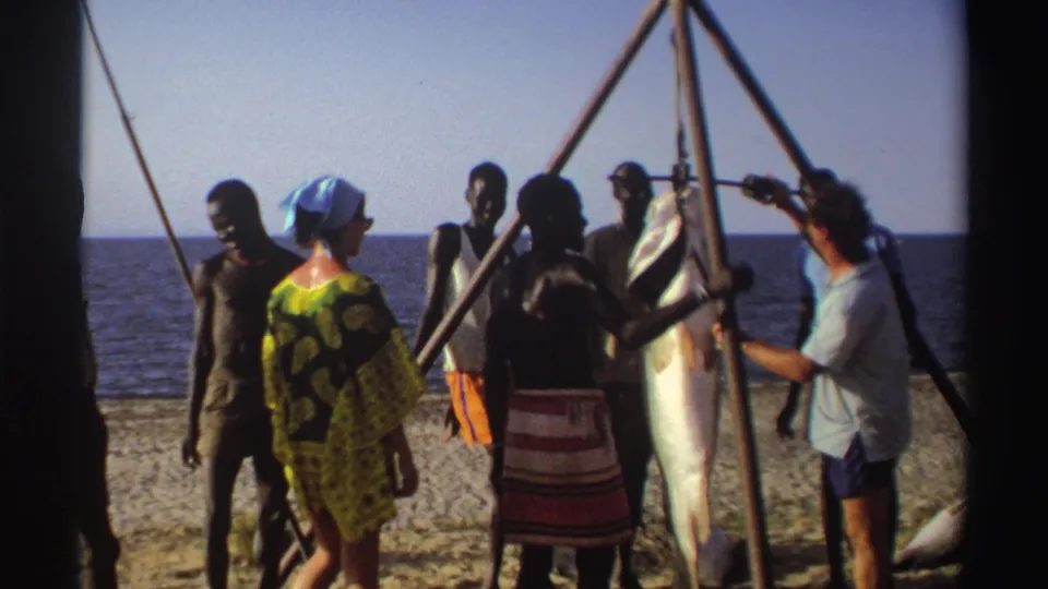 Adults in a 1969 Nigeria beach scene stand near a large catch suspended from a tripod, with the sea in the background.