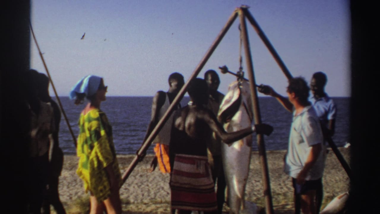 1969 Nigeria footage shows a woman in bright patterned clothing facing a group beside a large suspended fish on the beach.