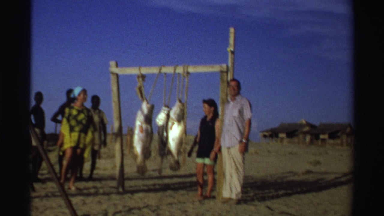 Tourists and local men stand beside large fish hanging from a wooden rack on a sandy beach.