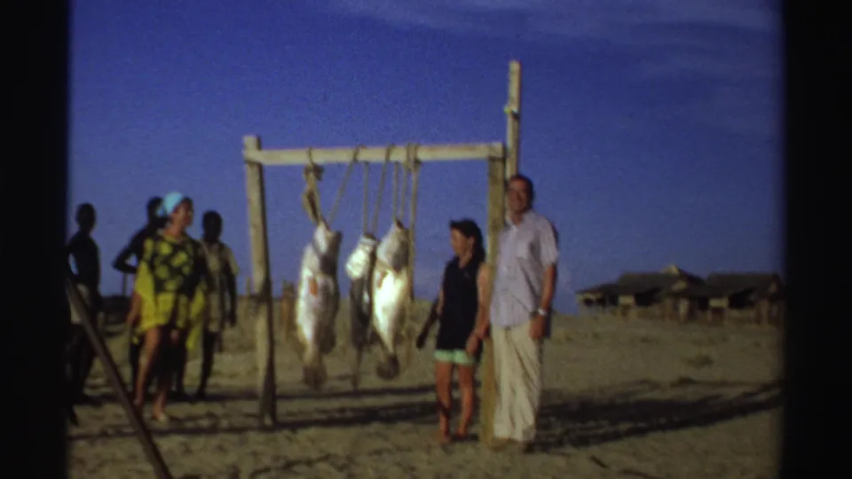 Nigeria 1969 home-movie frame showing tourists posing near large fish suspended from a wooden rack as local men stand nearby on the beach.