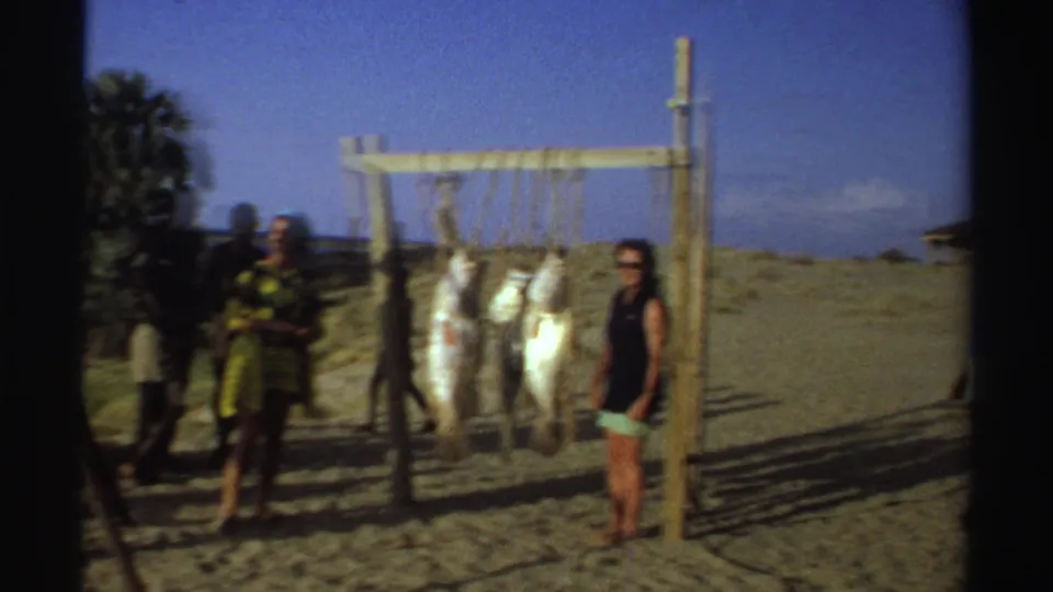 Nigeria 1969 beach frame with a visitor standing beside a wooden rack holding large fish while other people move nearby.