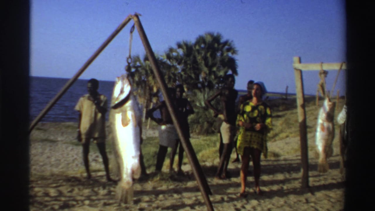 Nigeria 1969 home-movie frame showing a large fish suspended from a tripod pole near the ocean with tourists and local men nearby.
